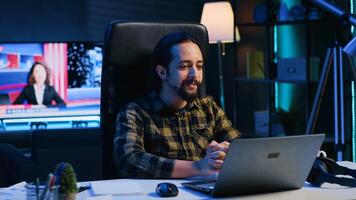 Smiling man relaxing in cozy living room by watching funny internet clips on laptop. Carefree caucasian person enjoying leisure time in RGB lit apartment, looking at online videos, camera A photo