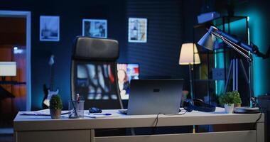 Focus on office desk in front of smart TV in blurry background displaying news program broadcasting. Laptop on table and television screen in empty room showing VOD channel, camera B photo