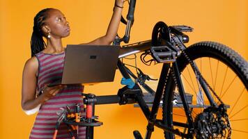 Technician with laptop in hand analyzing broken bike over studio background awaiting repair. Specialist using notebook to look for replacement parts for bicycle on repair stand, camera B photo