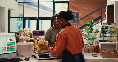 Merchant weighting bulk items on scale for regular client, selling various types on homemade products in reusable jars at checkout. Vendor chatting with buyer about healthy eating. Handheld shot. photo