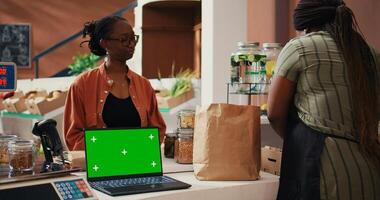 Retailer having laptop that shows greenscreen on display at cash register, giving eco products to client at market. Woman using computer with isolated chromakey template, mockup layout. Tripod shot. photo