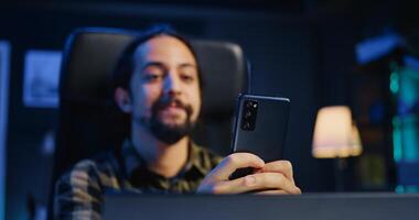 Happy man at home texting friend on cellphone, taking break from remote job in personal office. Joyful freelancer relaxing in apartment, talking with mate on internet using phone, camera B photo