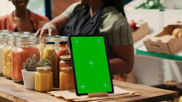 Tablet with greenscreen display placed on farmers market counter, next to homemade additives free sauces and cooking products in recyclable jars. Merchant displaying blank mockup template. photo