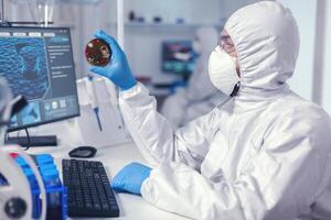 medicina investigador trabajos en SARS-CoV-2 en petri plato vestido en mono. doctores analizando vacuna evolución utilizando alto tecnología investigando diagnóstico en contra covid19 virus. foto