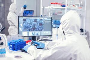 Chemist sits at his workplace in laboratory analyze coronavirus on computer dressed in ppe. Lab engineers conducting experiment for vaccine development against covid19 virus photo