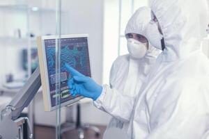 farmacia en proteccion traje que se discute acerca de cambios en vacuna composición en equipado laboratorio señalando escritorio. doctores examinando virus evolución utilizando alto tecnología investigando diagnóstico en contra covid19 foto