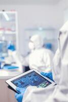 Chemist dressed in protection suit for coronavirus holding tablet pc in laboratory. Team of scientists conducting vaccine development using high tech technology for researching treatment against covid19 photo