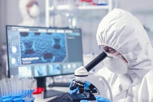 Woman in coverwall sitting at workplace in laboratory using microscope. Chemist researcher during global pandemic with covid-19 checking sample in biochemistry lab photo
