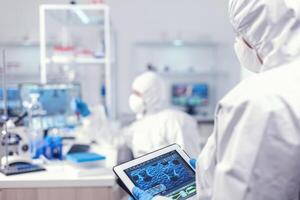 Doctor in ppe suit during coronavirus outbreak working in laboratory holding tablet pc. Team of scientists conducting vaccine development using high tech technology for researching treatment against covid19 photo