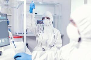 Lab scientist in healthcare lab holding micropipette with sample dressed in ppe suit against coronavirus. Team of microbiologists in research laboratory conducting experiment during global pandemic with covid19. photo