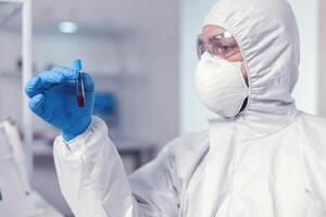 médico ingeniero mirando a muestra de sangre en tubo vistiendo proteccion traje. médico trabajando con varios bacterias y tejido, farmacéutico investigación para antibióticos en contra COVID-19. foto