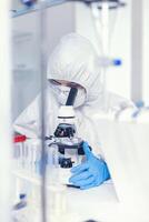 Scientist in sterile ppe suit checking sample of virus under microscope. Chemist in coverall examining vaccine evolution using high tech researching diagnosis against covid19 virus photo