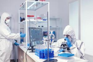 Close up of scientist using a microscope a microscope wearing ppe suit in equipped lab. Chemist researcher during global pandemic with covid-19 checking sample in biochemistry lab photo