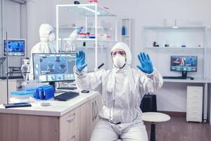 Virolog dressed in ppe suit developing coronavirus vaccine on digital screen making hand gesture. Medical scientist using modern technology to cure diseases and develop a cure. photo