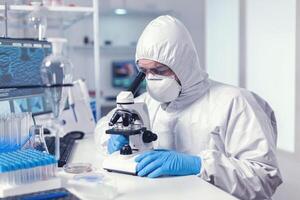 Doctor looking at blood sample with microscope wearing protection suit for covid19. Virolog in coverall during coronavirus outbreak conducting healthcare scientific analysis. photo