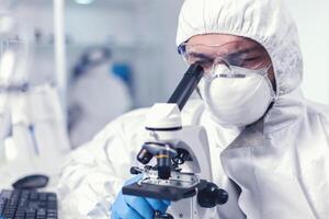 Chemical engineer wearing glasses conducting health investigation on microscope. Scientist in protective suit sitting at workplace using modern medical technology during global epidemic. photo