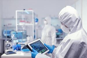 concentrado médico investigador utilizando digital tableta vestido en protector traje en contra infección con coronavirus. equipo de científicos conductible vacuna desarrollo utilizando alto tecnología tecnología para investigando tratamiento en contra covid19 foto