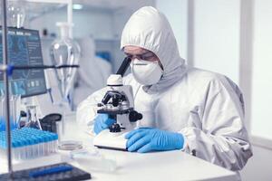 Microbiologist with protection glasses using microscope dressed ppe suit. Virolog in coverall during coronavirus outbreak conducting healthcare scientific analysis. photo