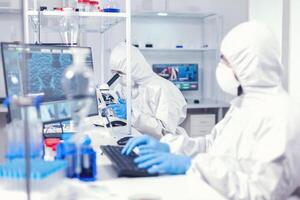mujer investigador que lleva fuera científico covid-19 prueba utilizando microscopio. médico ingeniero utilizando computadora durante global pandemia con coronavirus vestido en mono. foto