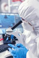 Medical scientist analysing sample on glass slide looking through microscope. Chemist researcher during global pandemic with covid-19 checking sample in biochemistry lab photo