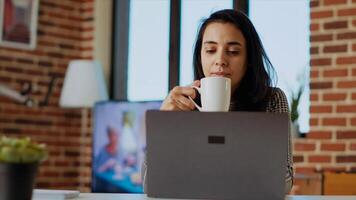 teletrabajador comprobación su correos electrónicos en ordenador portátil y disfrutando taza de café. remoto empleado trabajando desde hogar en brillante elegante acogedor Departamento personal oficina, Bebiendo caliente bebida, cámara si foto