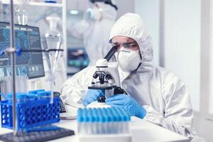 laboratorio técnico en ppe engranaje examinando virus muestras debajo microscopio en laboratorio. científico en protector traje sentado a lugar de trabajo utilizando moderno médico tecnología durante global epidemia. foto