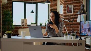 sonriente mujer avivado en haciendo en línea compras, cuidadosamente mecanografía débito tarjeta información en ordenador portátil teclado. persona agregando pago método en sitio web mientras en moderno Departamento vivo habitación, cámara un foto