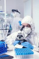 Chemist with face mask and ppe developing vaccine for covid looking through microscope. Scientist in protective suit sitting at workplace using modern medical technology during global epidemic. photo