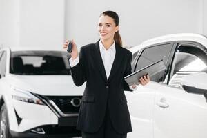 Female Competent car dealer in a suit with tablet at dealership center. Manager in auto dealership presents new cars. Dealer giving key to new owner in auto show or salon. photo