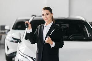 Female Competent car dealer in a suit with tablet at dealership center. Manager in auto dealership presents new cars. Dealer giving key to new owner in auto show or salon. photo