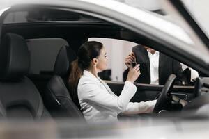 Car dealer giving keys to a young woman sitting inside the car. Joyful executive woman getting keys from new car. Happy auto owner buying transport at modern salon. photo