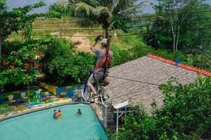 jepra jawa tengah, april 16, 2024 - rear view of a man on a bicycle designed to be a flying fox ride over a swimming pool surrounded by hills. photo