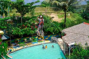 jepra jawa tengah, april 16, 2024 - rear view of a man on a bicycle designed to be a flying fox ride over a swimming pool surrounded by hills. photo