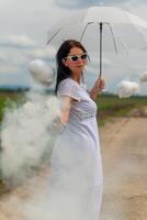 bonito mujer en un blanco vestir y linda nubes con fumar con un paraguas foto