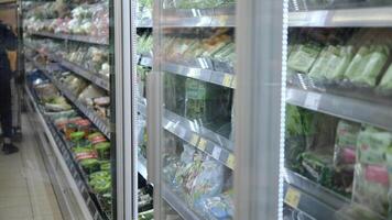Riga, Letland -28 april 2024 in een supermarkt, een vrouw bereikt in een koelkast, voorzichtig kiezen pakketjes van groenen. ze evalueert versheid van koelkast, trekken Groenen van koelkast video