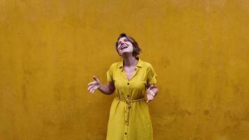 Cheerful young woman in a yellow dress laughing and gesturing against a textured mustard background, evoking summer vibes and International Day of Happiness video