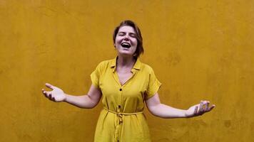 gioioso giovane caucasico donna nel un' giallo vestito ridendo con braccia disteso contro un' strutturato ocra parete, che rappresentano felicità e internazionale giorno di felicità video