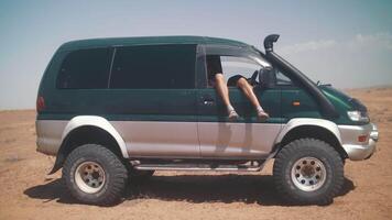 un' uomo è riposo con il suo gambe penzoloni su di il macchina. in giro il deserto video