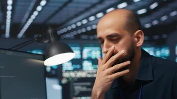 Overworked engineer feeling stressed while coding in data center facility, trying to ensure optimal performance of supercomputers. Tired man feeling fatigued while using PC to monitor server cabinets video