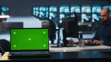 Focus on isolated screen laptop in data center with man in blurry background coding, managing server rows. Close up shot of chroma key notebook and IT expert overseeing rackmounts operating data video