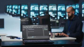 Focus on laptop used by programmer running code in blurry background for data center servers, troubleshooting rigs. Close up shot of code lines on notebook screen in server hub video