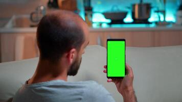 Caucasian male reading online social information on phone with mock up green screen chroma key display. Tired man sitting on sofa using isolated smartphone with technology internet video