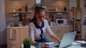 Hispanic lady holding credit card while doing online transaction using laptop in home kitchen late at night. Freelancer shopping online with electronic paymant on digital notebook conected to internet video