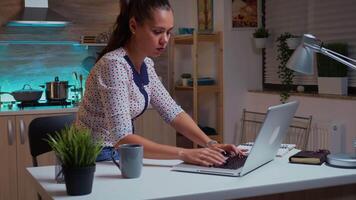 mulher verificação Assistir durante hora extra trabalhos dentro casa cozinha às meia noite fechamento computador portátil e saindo. ocupado focado empregado usando moderno tecnologia rede sem fio lendo digitando, procurando video