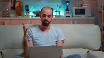 Caucasian male sitting on couch in pajamas working on laptop computer during videocall conference. Comfortable man relaxing on sofa chatting with business people late at night in kitchen video