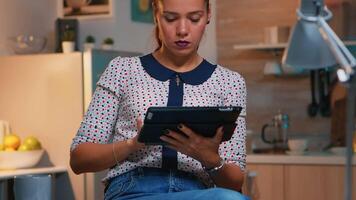 Businesswoman taking break using tablet late at night sitting in modern kitchen. Busy focused employee using modern technology network wireless doing overtime writing, searching. video