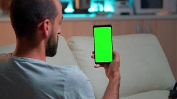 relaxado homem olhando às Smartphone com zombar acima verde tela croma chave exibição enquanto relaxante em sofá. caucasiano masculino segurando dentro horizontal modo telefone com isolado exibição atrasado às noite dentro cozinha video