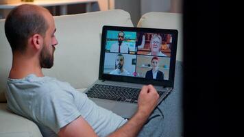 Over shoulder view of man in pajamas having online conversation with teammates working at online technology project using laptop computer, videoconference web internet communication video