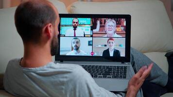 concentré adulte en utilisant portable ordinateur pendant appel vidéo conférence parlant à propos social affaires lien tandis que séance sur canapé. détendu homme travail à en ligne la communication projet heures supplémentaires dans cuisine video