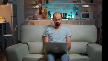 Focused man sitting on sofa and writing online project on laptop late at night in living room. Caucasian male working from home while watching tv, browsing on internet using network technology video